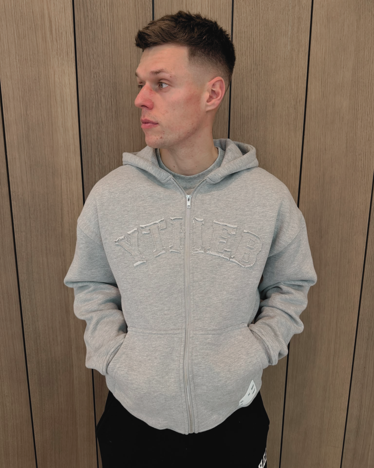 A man wearing a light gray double zip-up hoodie with 'YTHEB' embroidered across the chest, standing against a wood-paneled wall.