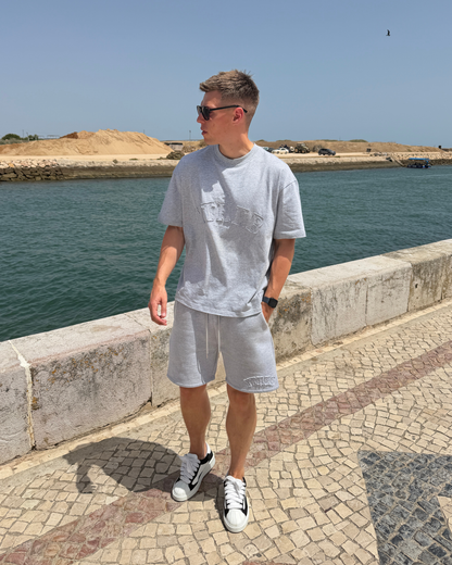 A man wearing a light gray graphic t-shirt, matching gray shorts, and white sneakers stands on a stone walkway next to the sea, looking to the side.