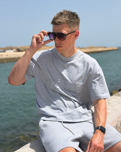 A man sitting outdoors by the water wearing a light gray heathered t-shirt and matching shorts, holding his sunglasses up to his eye.