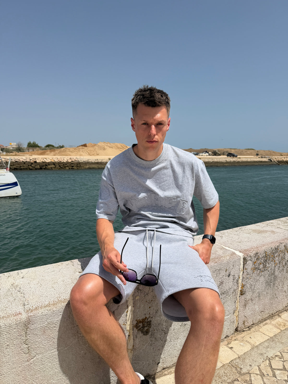 A man sitting on a stone wall by the sea, wearing a light gray crewneck t-shirt and matching light gray shorts, holding a pair of sunglasses.