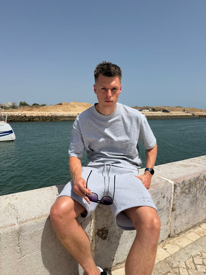 A man sitting on a stone wall by the sea, wearing a light gray crewneck t-shirt and matching light gray shorts, holding a pair of sunglasses.