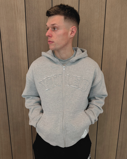A man wearing a light gray double zip-up hoodie with 'YTHEB' embroidered across the chest, standing against a wood-paneled wall.