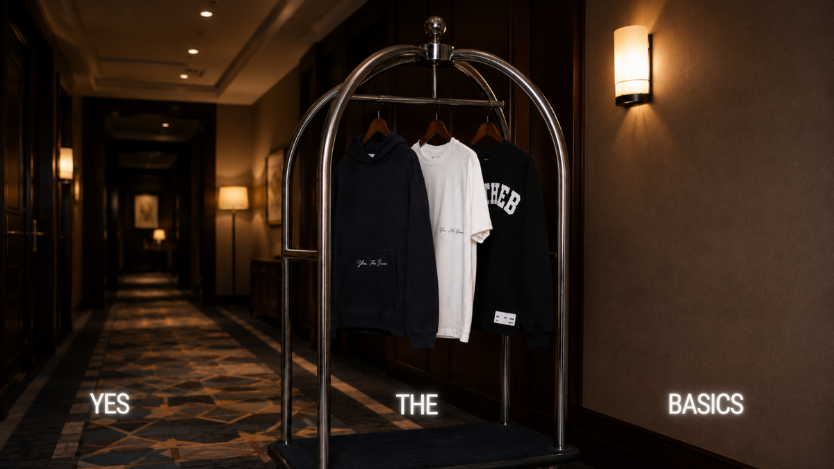 Navy hoodie, white tee and black YTHEB long sleeve hanging on a chrome rail in a hotel corridor