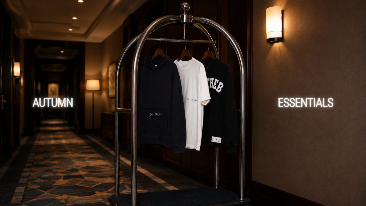 A metal garment cart in a hotel hallway displays three hoodies: a navy one, a white one, and a black one with 'ESSEBR' lettering. The hallway has warm lighting, patterned carpet, and wall-mounted lamps, with the words 'AUTUMN' and 'ESSENTIALS' projected on the walls.